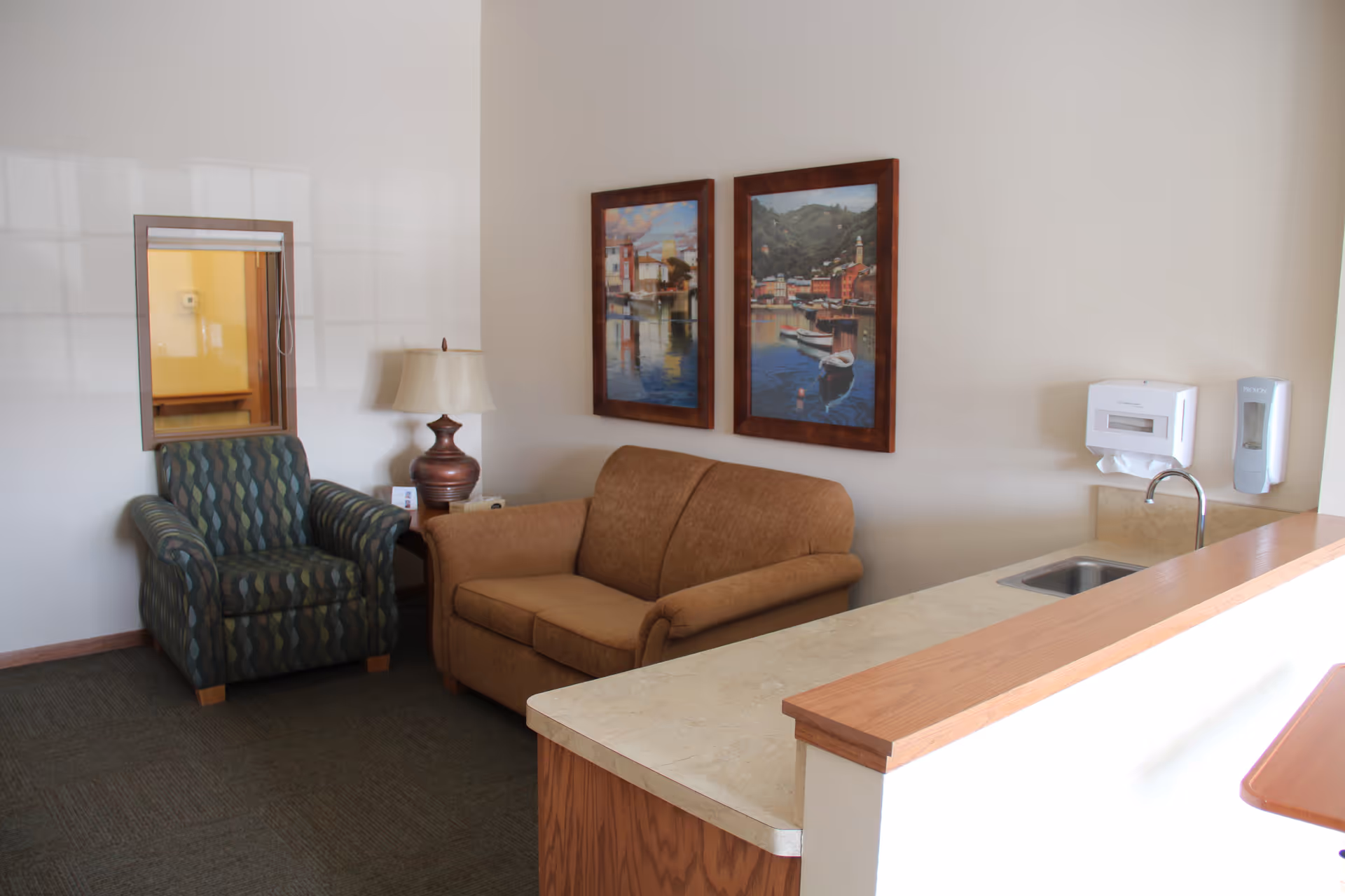 A cozy senior living living room area with a tan sofa, a patterned armchair, two framed paintings, a lamp, and a kitchenette counter with a sink.