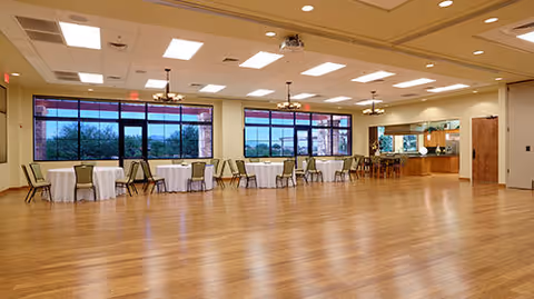 A spacious dining room with wooden flooring, several round tables covered with white tablecloths, and green chairs arranged around them. Large windows along one wall provide natural light and a view of trees outside. Ceiling lights and fans are evenly spaced across the ceiling. A serving area or kitchen pass-through window is visible in the background.