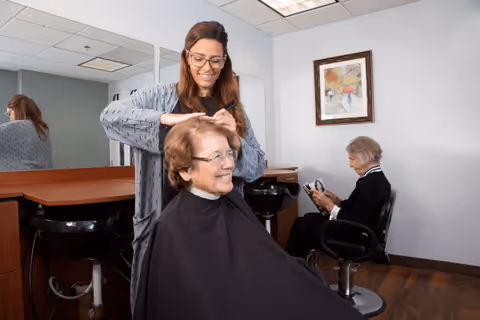 A hairdresser is cutting the hair of a smiling elderly woman seated in a salon chair. Another elderly woman is seated in the background, reading a magazine. The room has mirrors, salon chairs, and a framed painting on the wall.