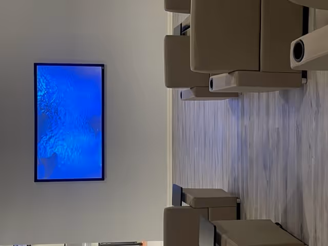 A lounge with beige armchairs and ottomans on wood floors facing a wall-mounted television showing a blue screen.