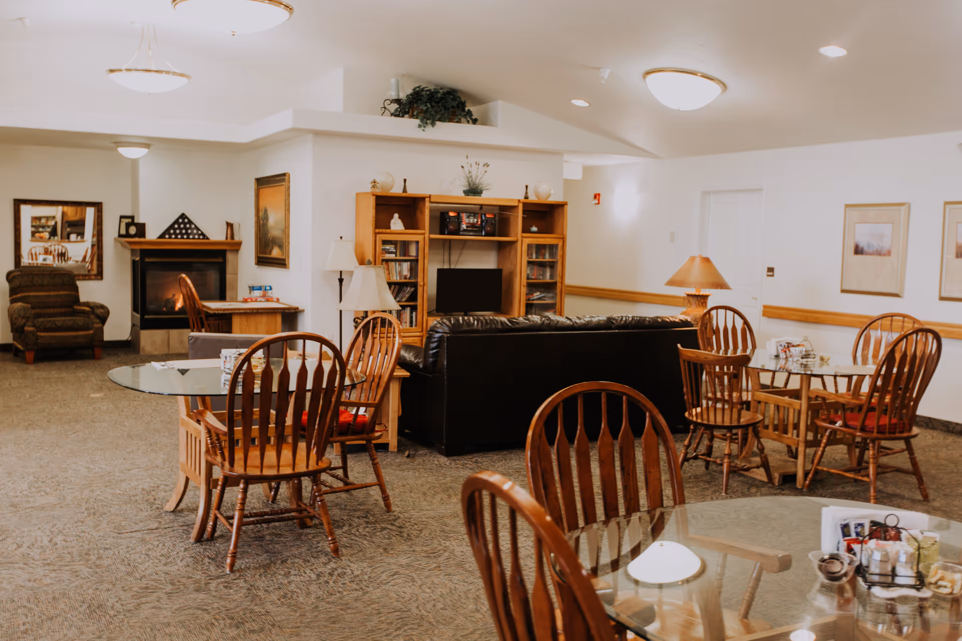 Bright communal lounge with wooden tables and chairs, a black leather sofa facing a bookcase and TV, and a fireplace.