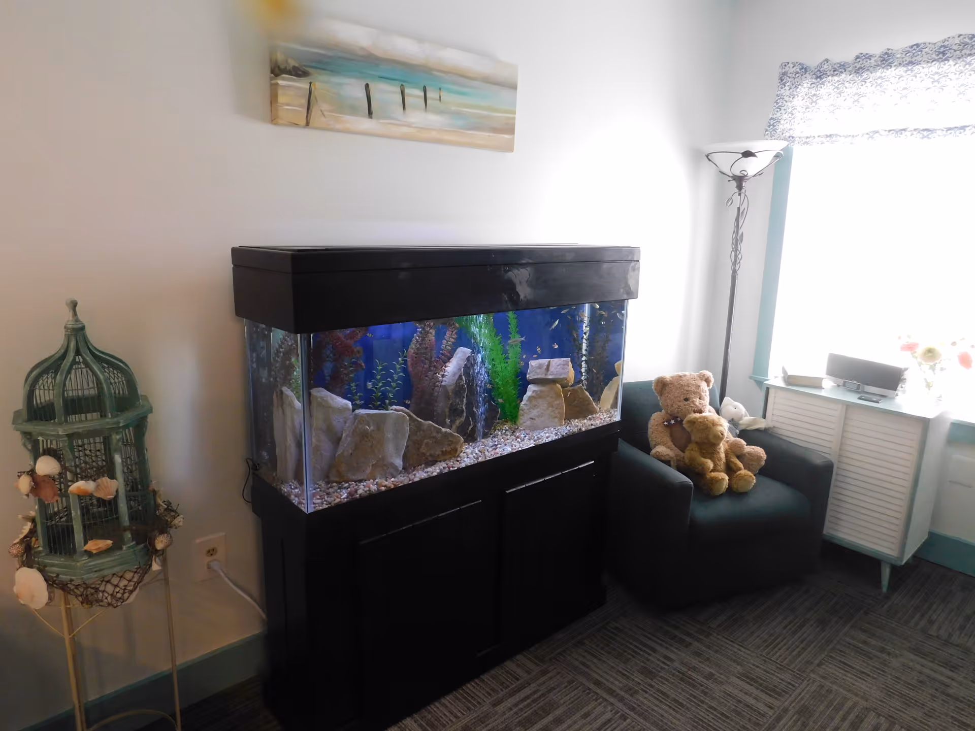 A cozy corner in a senior living community room featuring a large fish tank with rocks and plants, a dark green armchair with three teddy bears sitting on it, a tall floor lamp, a small cabinet with a speaker and flowers on top, and a decorative birdcage stand with seashells. A painting of a beach scene hangs on the wall above the fish tank.