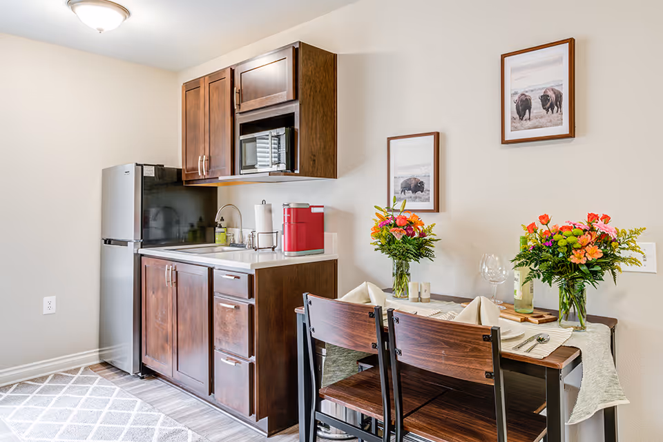 A small kitchen area with dark wooden cabinets, a stainless steel refrigerator, a microwave, and a sink. Next to the kitchen is a dining table set for two with folded napkins, wine glasses, and two vases of colorful flowers. Two framed pictures of bison hang on the wall above the table.
