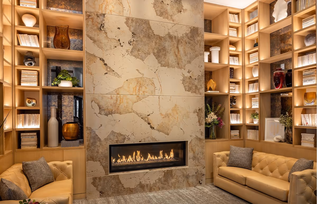 A cozy living room with a modern rectangular fireplace set in a large stone wall. The room features built-in wooden shelves filled with books, decorative vases, and plants. Two beige tufted leather sofas with gray cushions face each other across a textured rug.