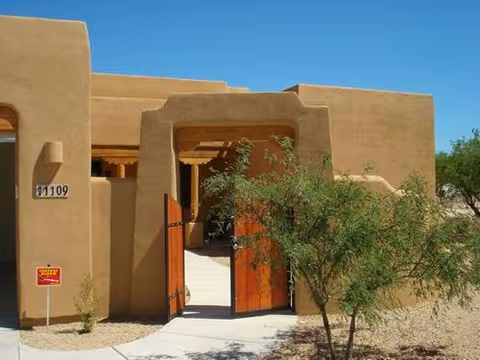 Exterior view of a southwestern-style building with tan stucco walls, a wooden gate partially open, a small tree in front, and a clear blue sky.