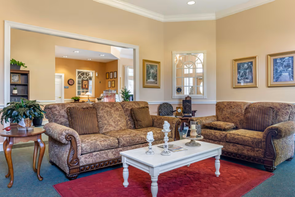 A cozy living room with two patterned brown sofas, a white coffee table with decorative items, a red rug, and framed pictures on beige walls. There is a small wooden side table with a plant on it and a mirror on the wall reflecting part of the room.