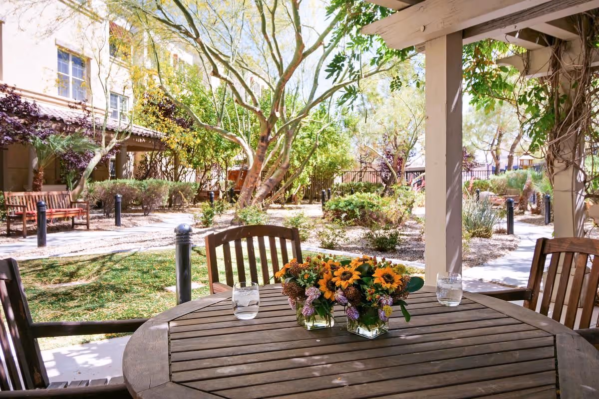 Wooden patio table with vases of flowers and glasses under a pergola overlooking a landscaped courtyard with benches and trees.