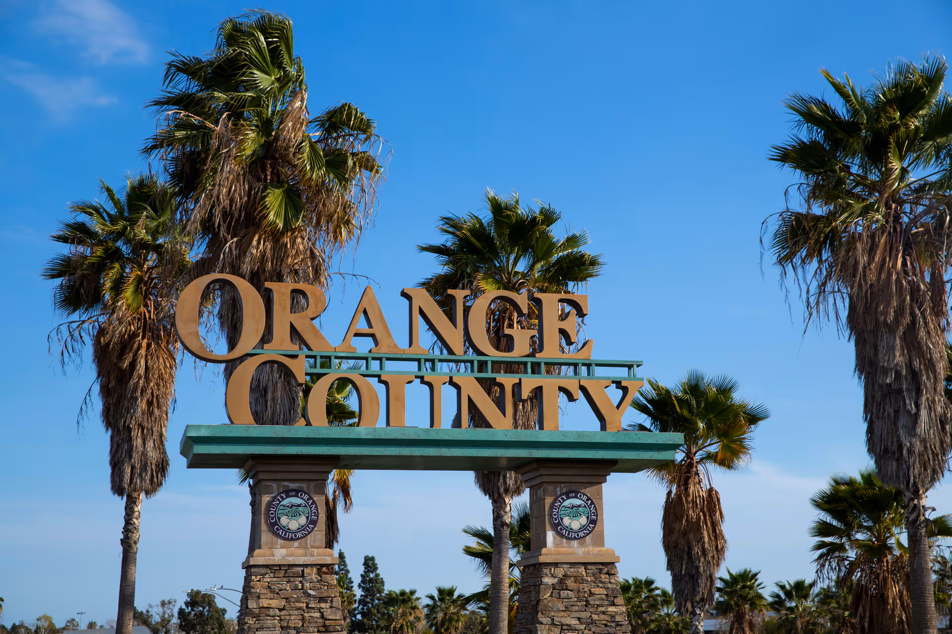 Large stone and metal sign reading 'ORANGE COUNTY' with palm trees and a clear blue sky in the background.