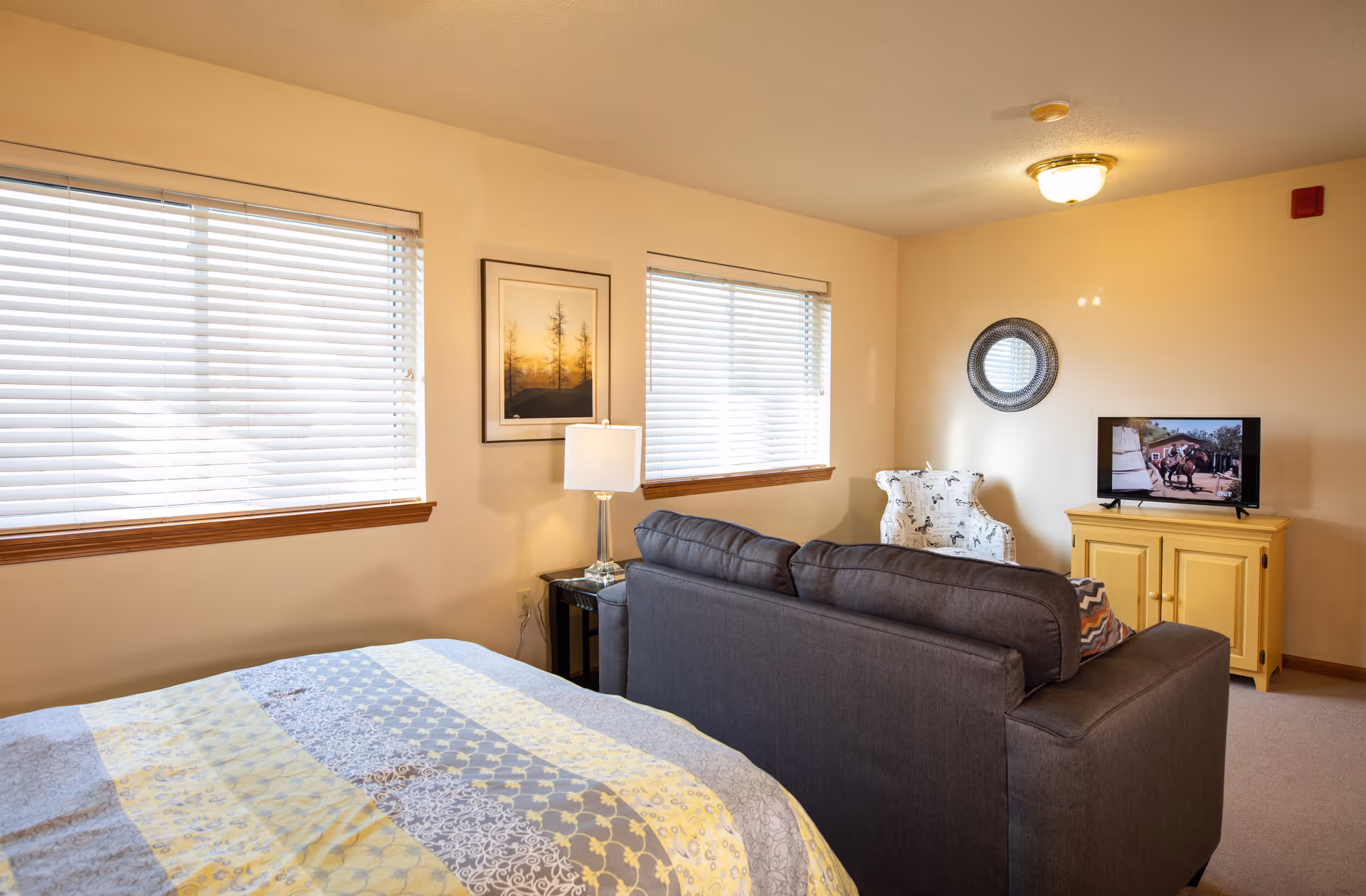 A cozy living space featuring a bed with a patterned quilt, a dark gray sofa facing a yellow TV stand with a flat-screen television, a white armchair with a black and white pattern, two windows with blinds, a framed picture on the wall, a table lamp on a small side table, and a round decorative mirror on the wall.