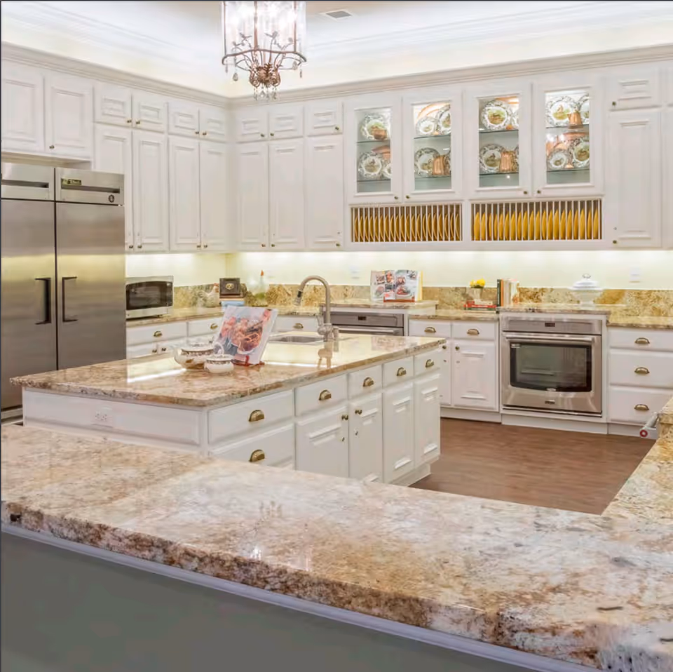 A spacious, well-lit kitchen with white cabinetry, granite countertops, and stainless steel appliances including a double-door refrigerator and built-in oven. The kitchen features a large island with a sink and decorative items, glass-front cabinets displaying plates, and a chandelier hanging from the ceiling.