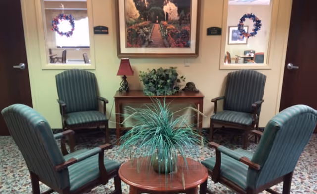 A small seating area with four green striped armchairs arranged around a round wooden table with a potted plant. Behind the chairs is a wooden console table with a red lamp, a green plant, and a decorative item. Above the console table hangs a framed picture of a garden pathway. Two doors and two windows with wreaths are visible in the background.