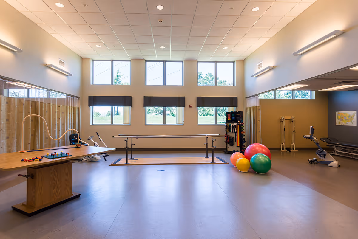 A spacious physical therapy or rehabilitation room with large windows letting in natural light. The room contains parallel bars for walking exercises, colorful exercise balls, a stationary bike, a therapy table with various therapy tools, and a rack with small weights. The walls are light-colored, and the ceiling has recessed lighting.