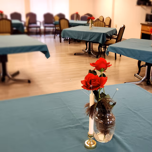 Dining room with teal-covered tables and a vase of red roses and a candle as a centerpiece in the foreground.