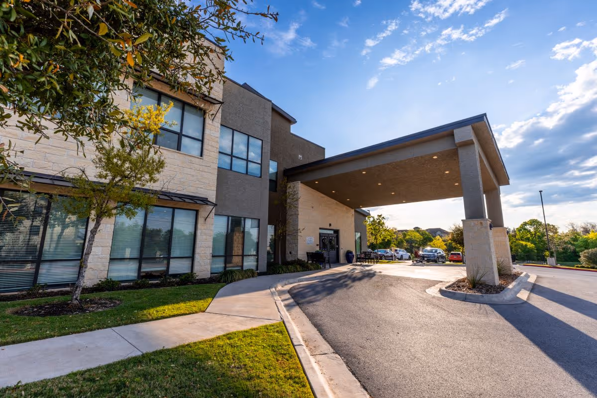Exterior view of Tech Ridge Oaks facility showing a modern two-story building with large windows, a covered entrance driveway, landscaped greenery, and a few parked cars under a partly cloudy sky.