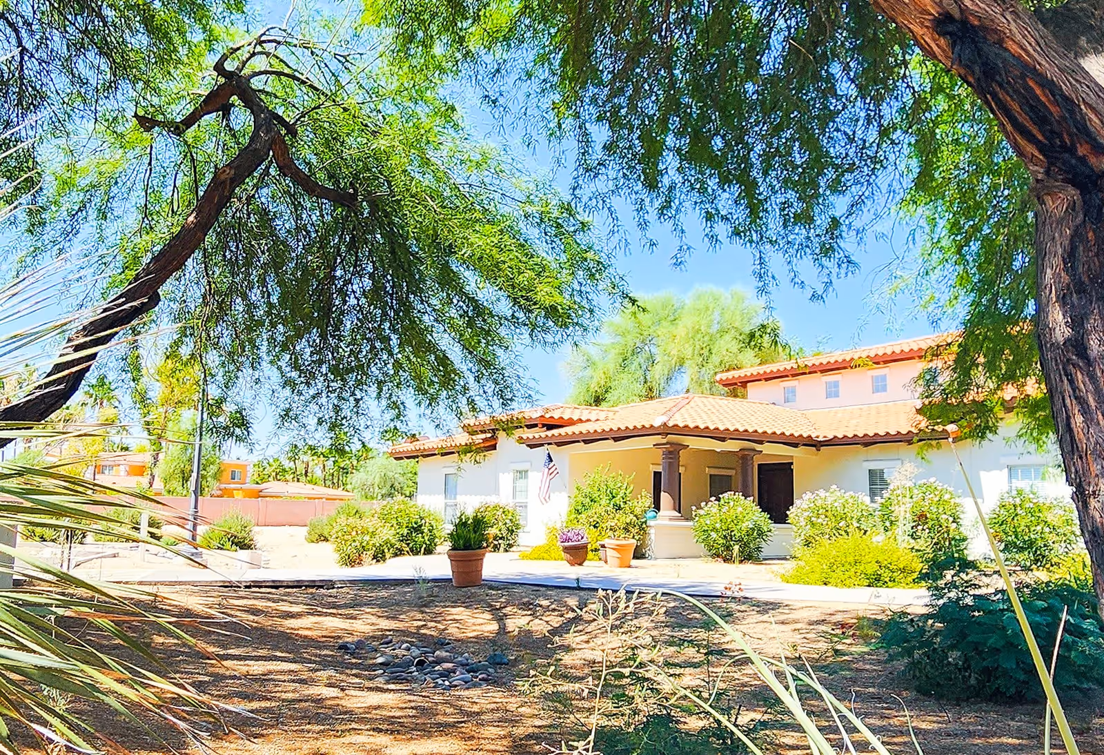 Front exterior of a single-story stucco building with a red tile roof, surrounded by trees, shrubs, potted plants, and a shaded walkway.
