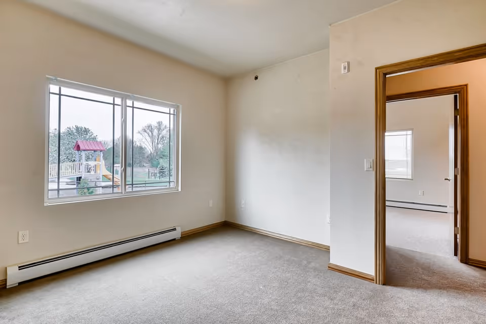 Empty carpeted room with a large window overlooking an outdoor playground and an open doorway to another room.