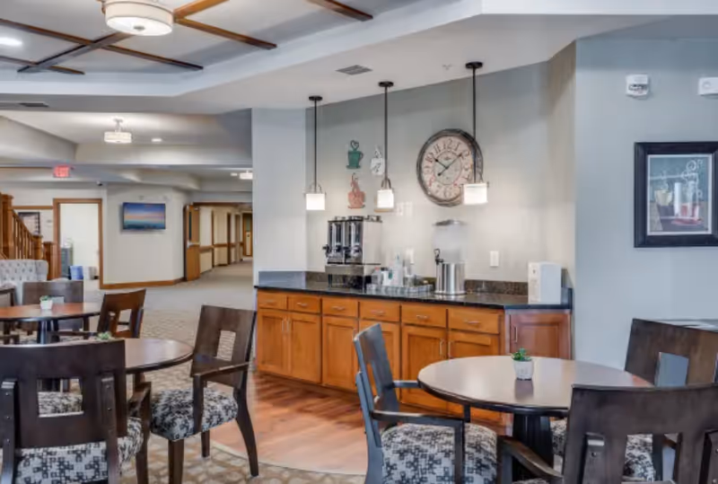 Interior view of a senior living facility common area with round tables and chairs, a coffee and beverage station with wooden cabinets, pendant lights, a large wall clock, and framed artwork on the wall.