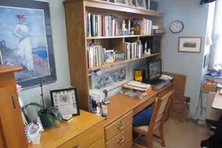 A wooden desk with a bookshelf above, computer monitor, and papers in a cozy study room.