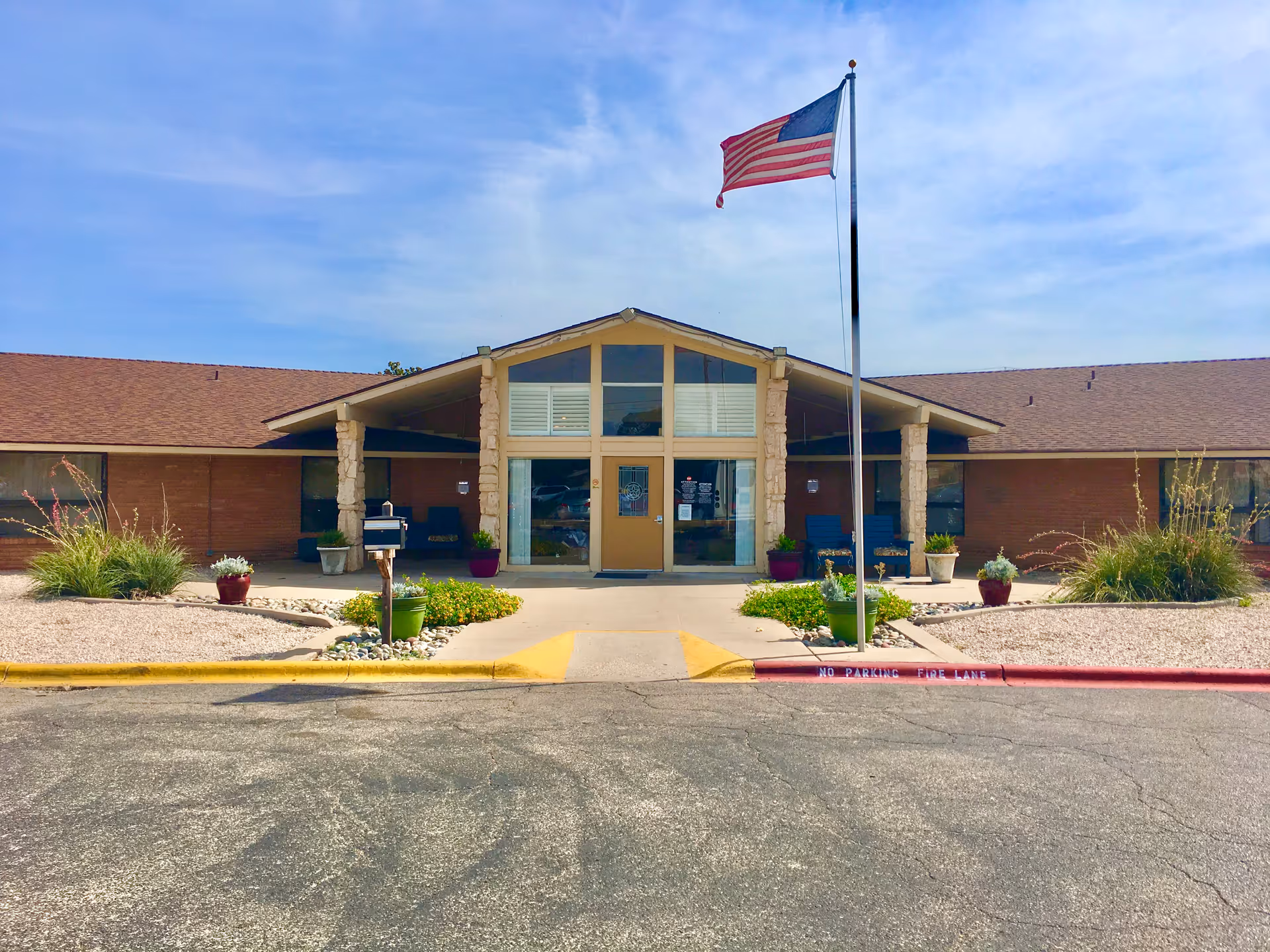 Front exterior view of Buena Vida Nursing and Rehabilitation Odessa building with a flagpole flying the American flag, a paved driveway, and landscaped areas with plants and flowers.