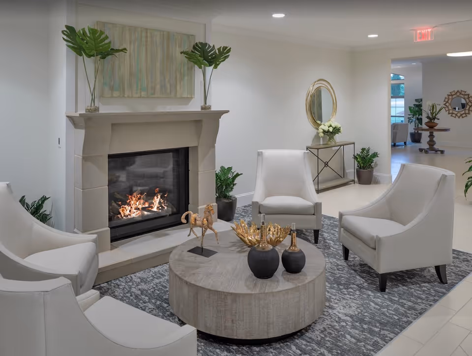 Cozy lounge with four white armchairs around a round wooden coffee table, decorative vases and a lit fireplace against a white wall.