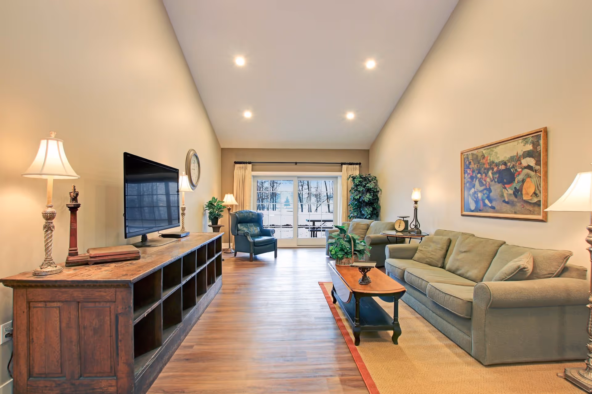 A spacious living room with wooden flooring, a large green sofa, a wooden coffee table with a plant, a green armchair near a window with curtains, a wooden TV stand with a flat-screen TV, lamps, and a framed painting on the wall.