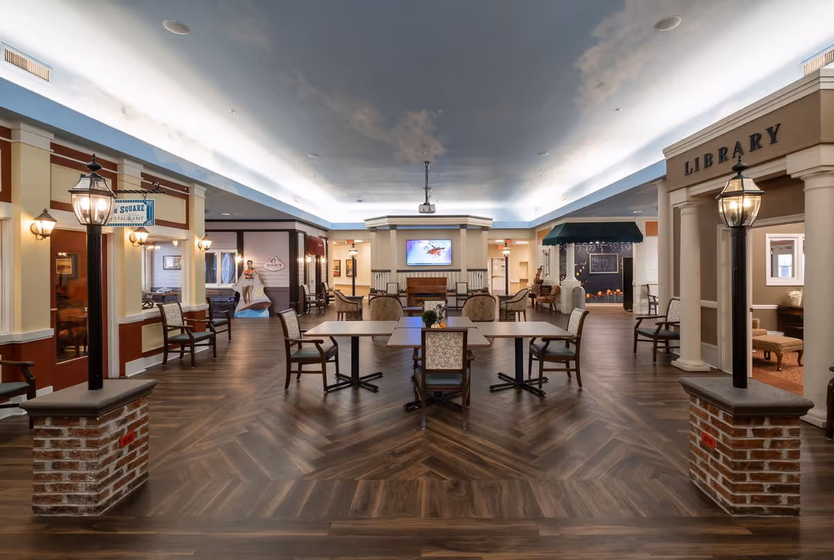Interior view of a senior living facility common area with wooden flooring and a ceiling painted to resemble the sky with clouds. There are tables and chairs arranged in the center, a fireplace with a TV above it at the far end, and signs indicating a library on the right and a restaurant on the left. The space is well-lit with decorative streetlamp-style lights and wall sconces.