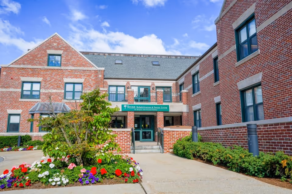 Exterior view of Stone Rehabilitation and Senior Living building with red brick walls, green framed windows, a green entrance door, and a garden with colorful flowers and shrubs in front under a partly cloudy blue sky.