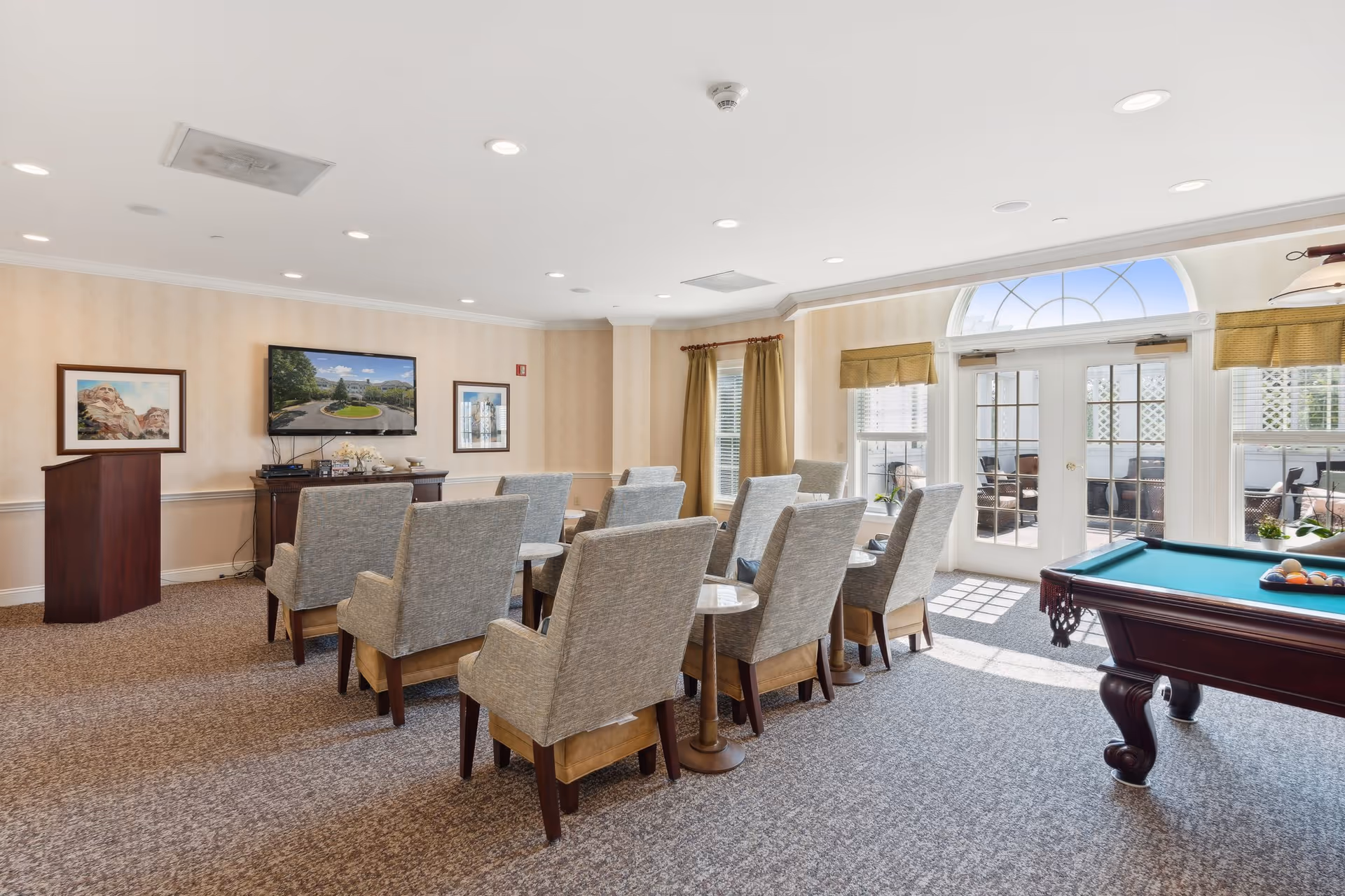 A bright and spacious common area with rows of cushioned chairs facing a wall-mounted TV. The room features beige walls, carpeted floors, framed artwork, and large windows with curtains letting in natural light. A pool table is positioned on the right side near glass doors leading to an outdoor patio.