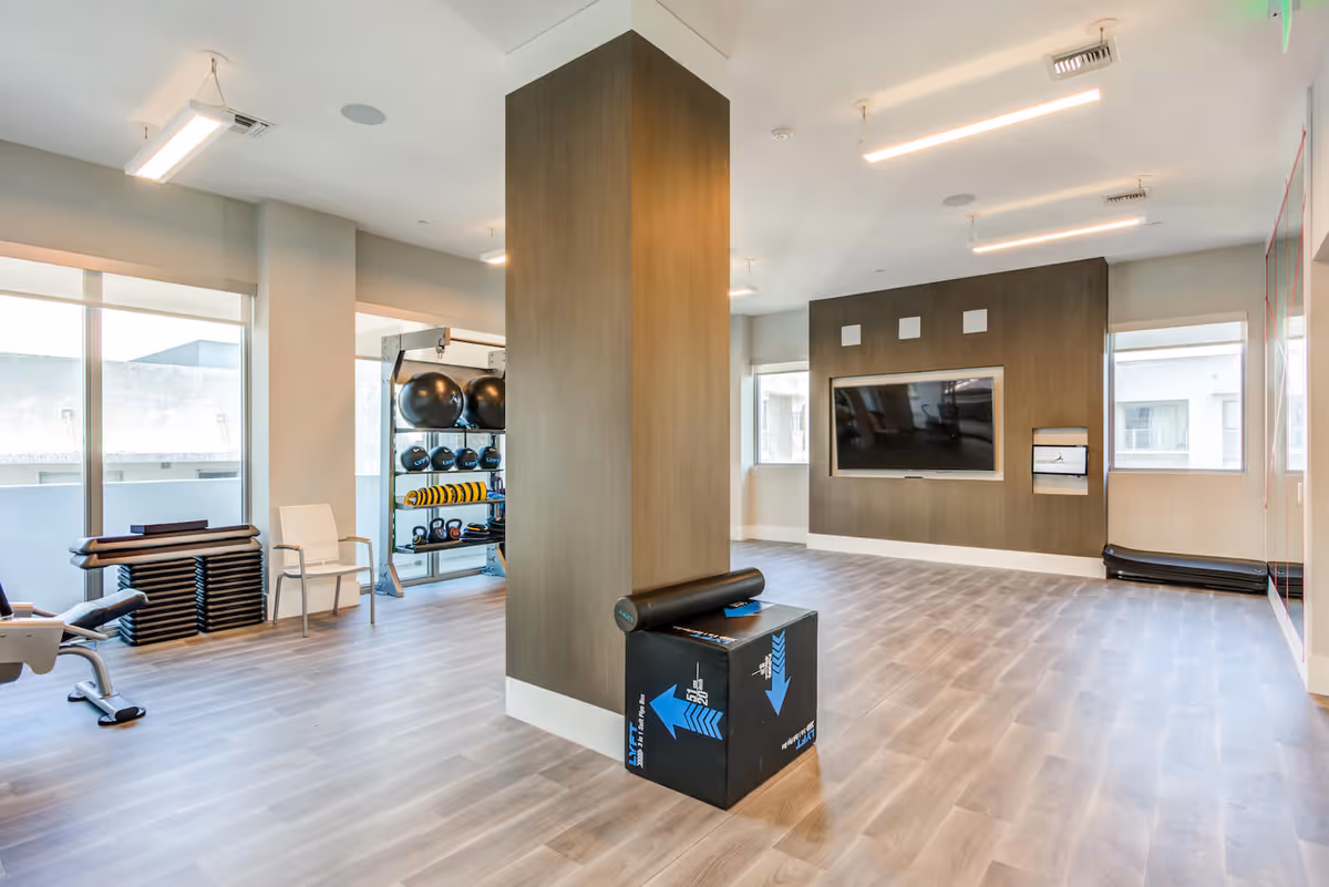 A modern fitness room with light wood flooring, large windows, and a central column. The room contains exercise equipment including a bench, stacked aerobic steps, kettlebells, exercise balls, and a foam roller on a plyometric box. A large flat-screen TV is mounted on a dark wood-paneled wall, and there is a smaller screen below it. The space is well-lit with ceiling lights and has a clean, open layout.
