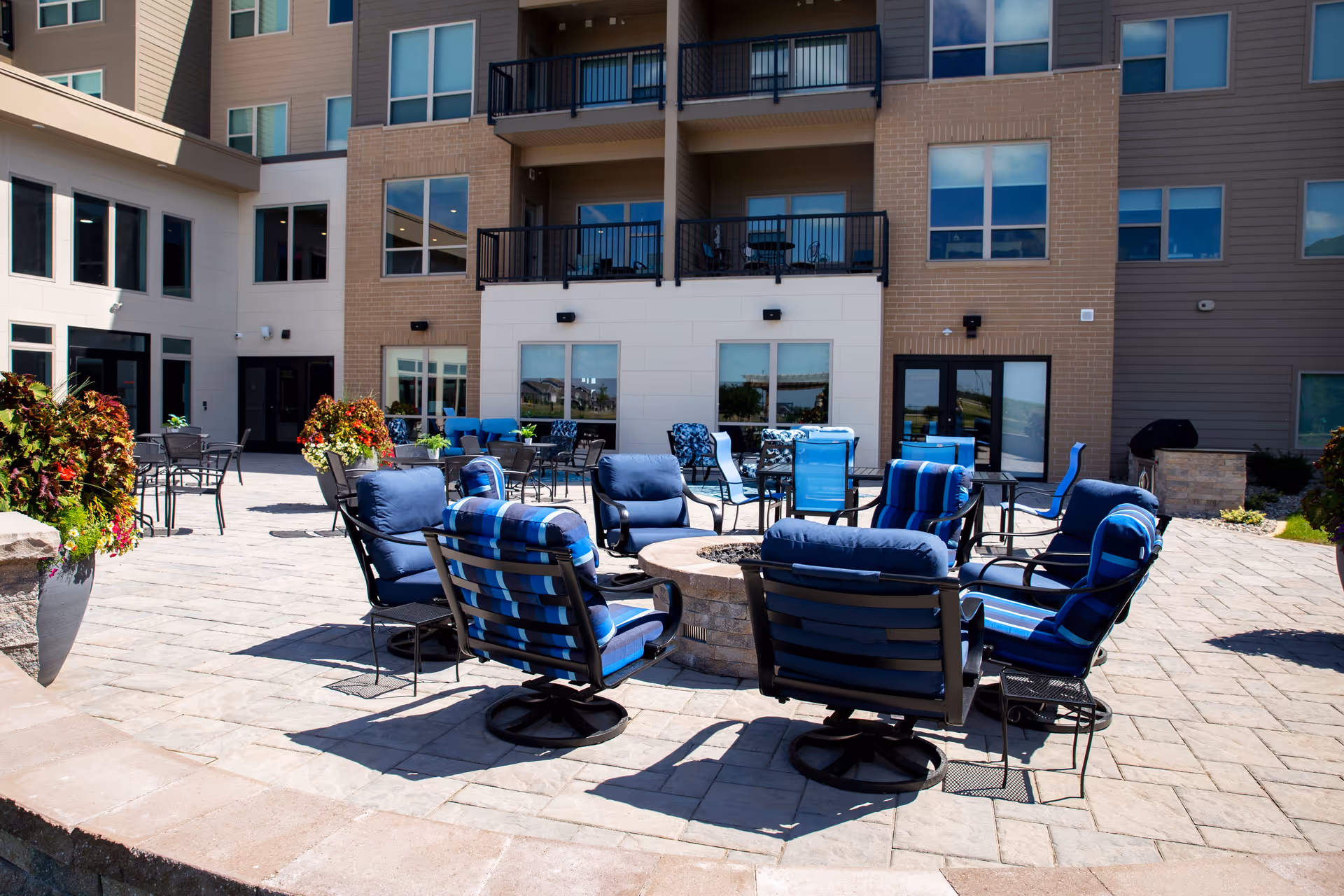 Outdoor patio area at Attivo Trail Waukee with cushioned blue chairs arranged around a circular fire pit, additional tables and chairs in the background, and a multi-story building with balconies and large windows.