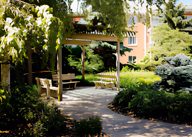 A shaded outdoor seating area with benches under a wooden pergola surrounded by lush green trees and bushes, with a building visible in the background.