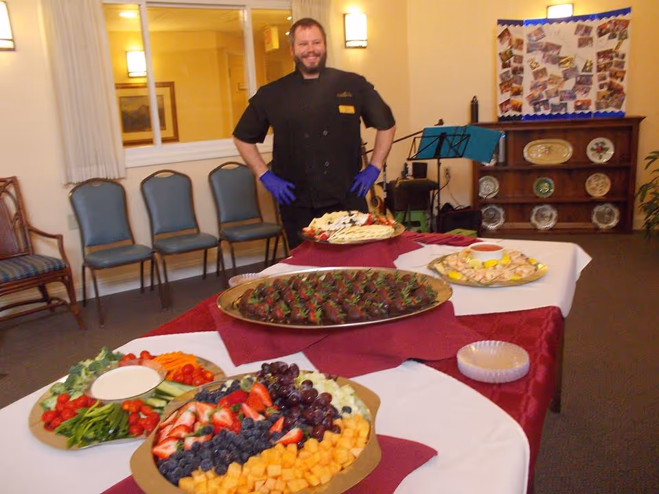 A smiling staff member stands behind a buffet table filled with platters of fruit, vegetables, and appetizers in a dining/event room.