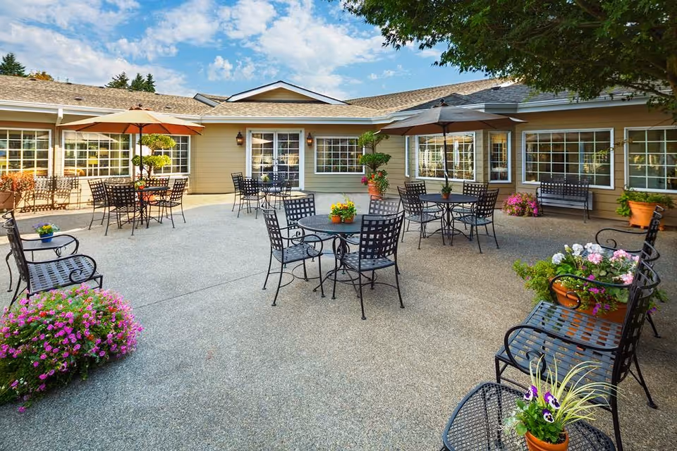 Outdoor patio area at Canterbury Inn Assisted Living Community with multiple black metal tables and chairs, some shaded by umbrellas. The patio is decorated with potted flowers and plants, surrounded by the building's beige exterior walls and large windows under a partly cloudy sky.