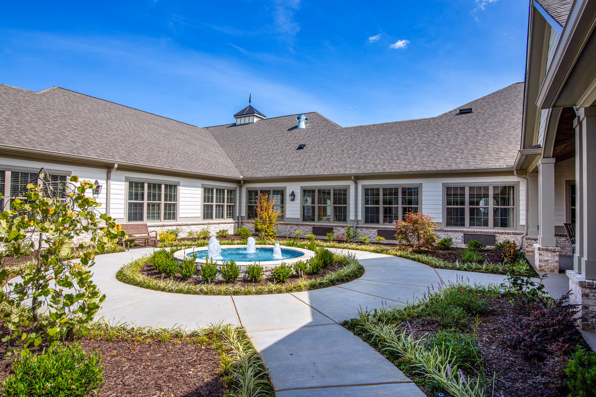 Outdoor courtyard area at Orchard at Athens featuring a circular water fountain in the center surrounded by a concrete walkway and landscaped garden beds, with a single-story building with multiple windows and a gray shingled roof in the background under a clear blue sky.