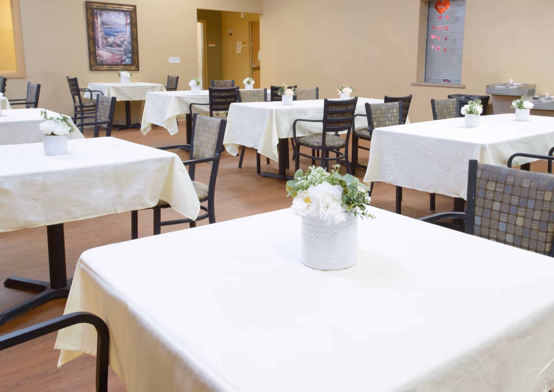 A dining room in a senior living facility with multiple tables covered in white tablecloths, each adorned with a small white flower arrangement. The room has beige walls, wooden flooring, and several chairs around the tables. A framed painting is visible on one wall, and there are water fountains and a window with heart decorations in the background.