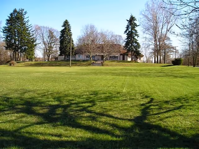 A single-story building surrounded by a large, well-maintained grassy lawn with several tall trees around it under a clear blue sky.