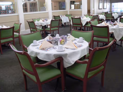A dining room with multiple round tables covered with white tablecloths, each set with folded napkins, cups, saucers, and silverware. The chairs around the tables have wooden frames with green cushions. The room has beige walls with windows and partitions.