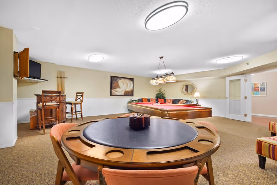 A recreational room with a round wooden game table with cup holders and four chairs in the foreground, a pool table with a red felt surface and hanging lights above it in the background, a small bar area with two high chairs and a wall-mounted TV on the left, and a seating area with a couch and colorful cushions along the back wall. The room has beige walls, carpeted floor, and ceiling lights.