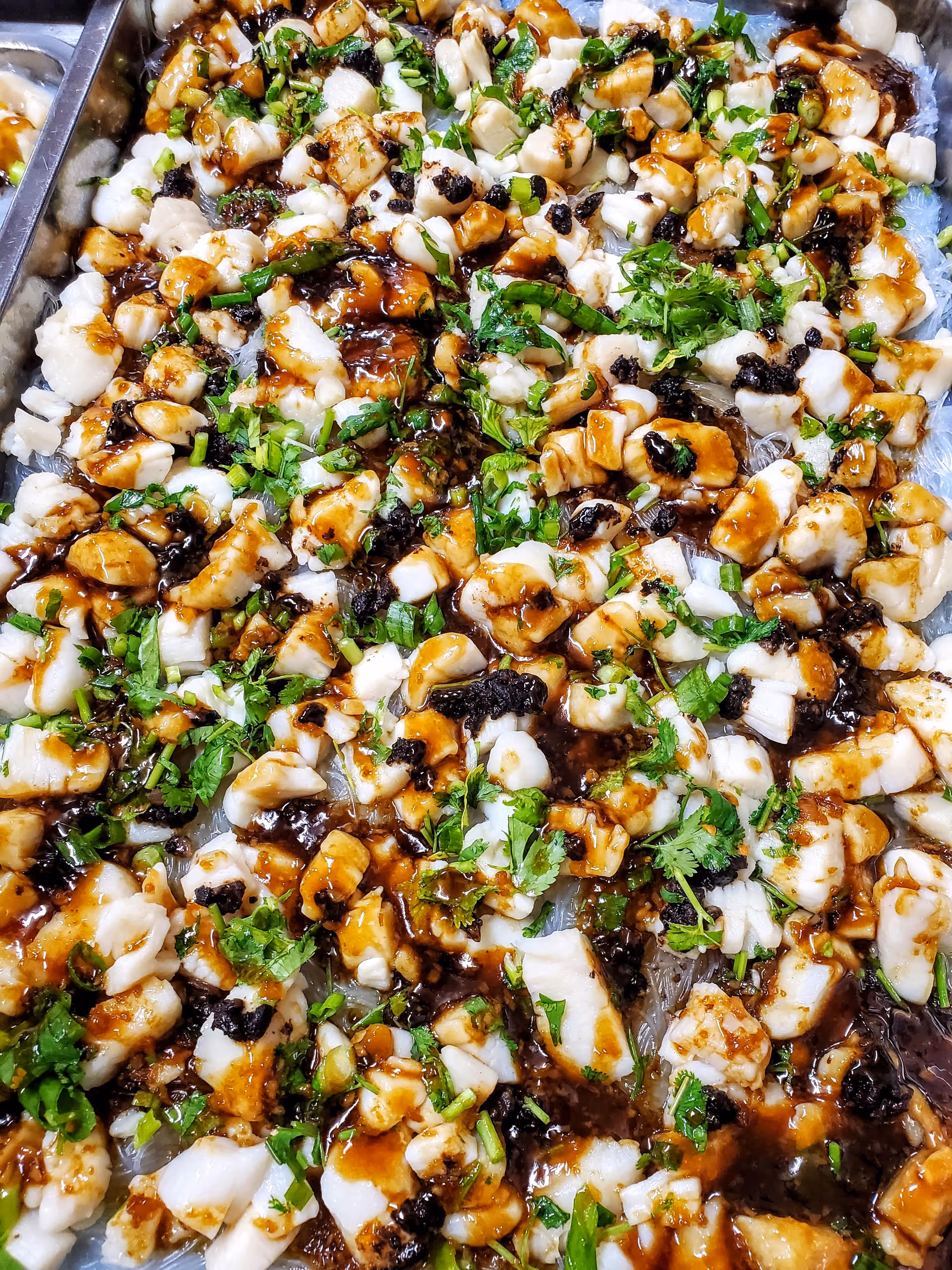 Close-up view of a dish featuring chopped white seafood pieces, garnished with chopped green herbs and drizzled with a dark sauce.