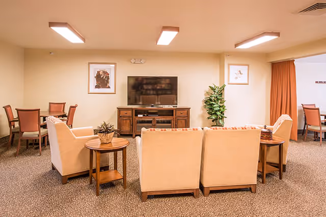 A cozy living room area with beige armchairs and a sofa arranged around a wooden TV stand with a flat-screen television. There are side tables with plants, framed artwork on the walls, and a dining area with chairs visible in the background.