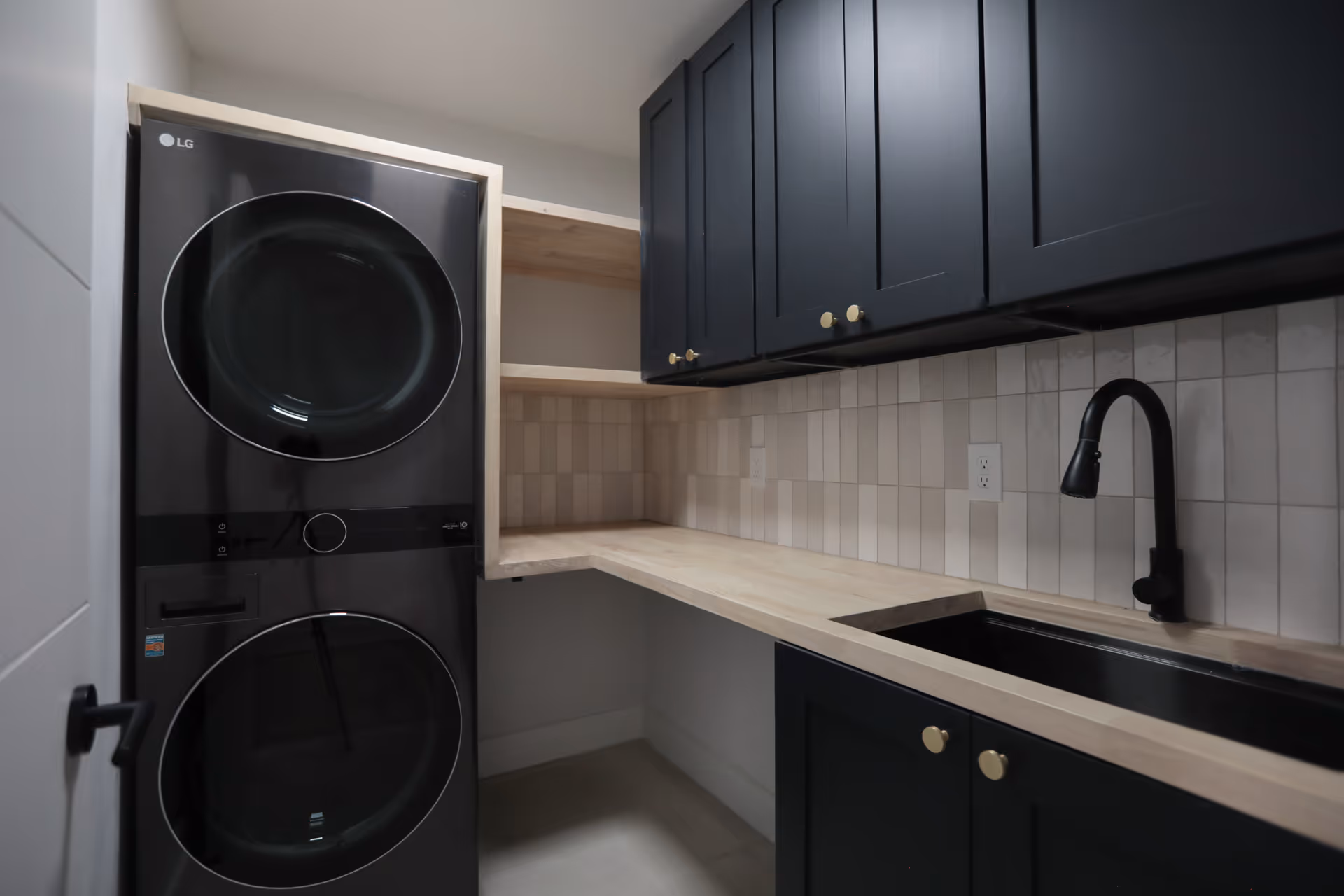 Modern laundry room with stacked black LG washer and dryer on the left, light wood countertops, black sink with a black faucet, beige tiled backsplash, and dark blue cabinets with gold knobs.