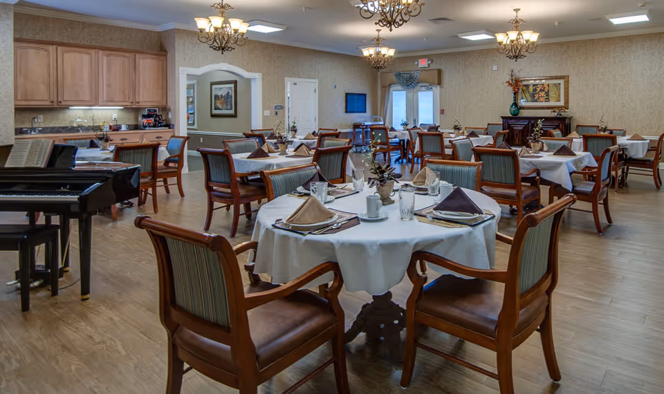 A spacious dining room in a senior living facility with multiple round tables covered with white tablecloths, each set with napkins, glasses, and silverware. Wooden chairs with striped upholstery surround the tables. A piano is visible on the left side near a small kitchen area with wooden cabinets. The room is well-lit with chandeliers and has a warm, inviting atmosphere.