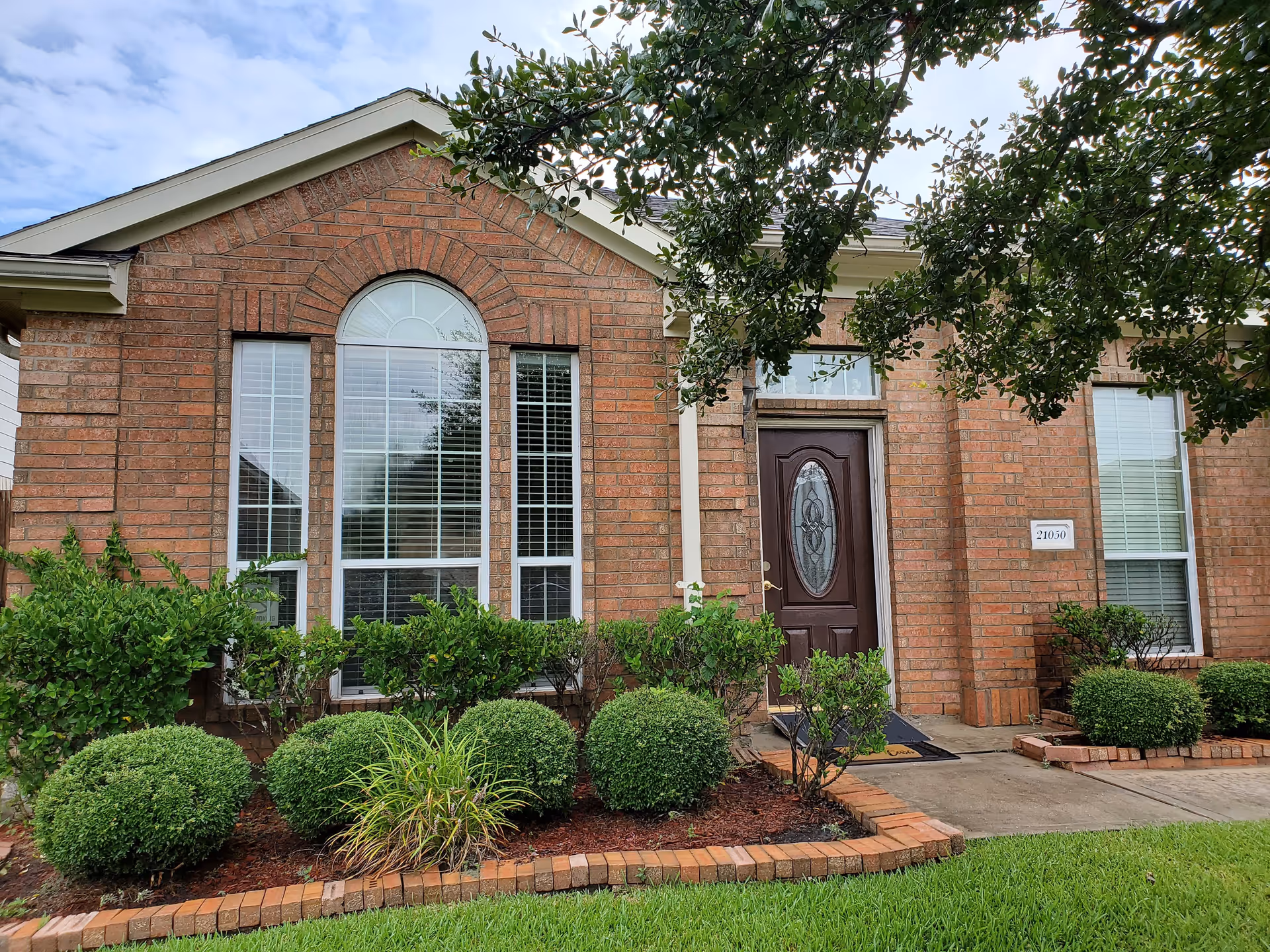 Front exterior view of a brick building with a dark wooden door featuring an oval glass design. The building has three large windows with white blinds and is surrounded by neatly trimmed bushes and a green lawn. A tree branch partially covers the top right corner of the image.