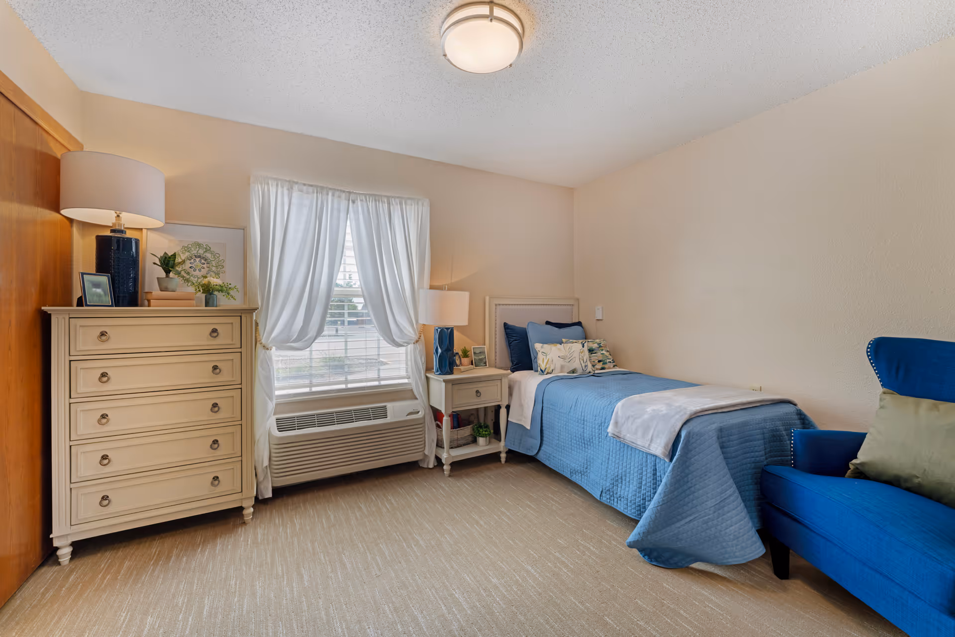 Well-lit bedroom with a single bed in blue linens, a dresser and nightstand by a curtained window, and a blue armchair.