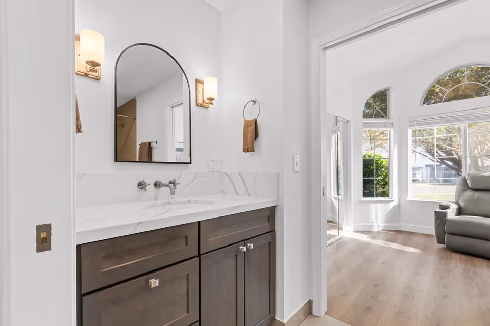 Modern bathroom vanity with a marble countertop, arched mirror and wall sconces opening into a sunlit living area with large windows and a recliner.