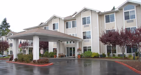 Exterior view of a three-story senior living facility named Brookdale Allenmore with a covered entrance, landscaped bushes and trees, and a wet driveway.