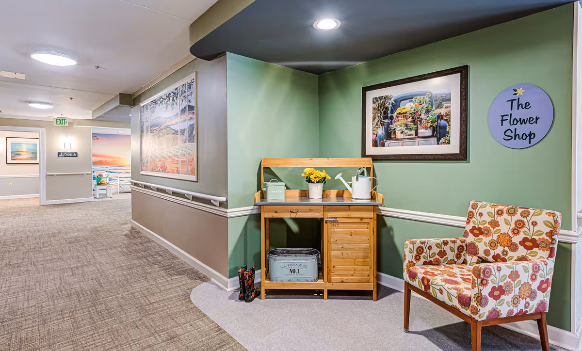 A hallway corner in a senior living facility with green walls and beige carpet. There is a wooden potting bench with a watering can, a small container labeled 'seeds', and a pot of yellow flowers. Next to the bench is a floral patterned armchair. On the wall above the bench is a framed picture of a truck bed filled with flowers and a round sign that reads 'The Flower Shop'. The hallway extends to the left with more artwork and an exit sign visible.
