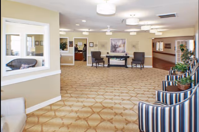 A spacious interior common area with beige walls and patterned carpet. The room features several chairs, including striped armchairs on the right and two dark armchairs near a table with decorative items. There are framed artworks on the walls and ceiling lights providing illumination. In the background, two people are seen standing near a desk or reception area.