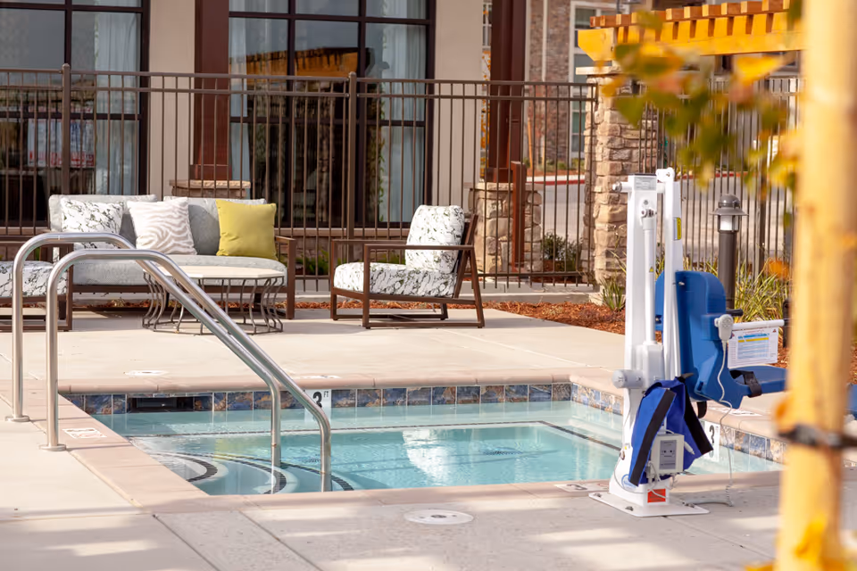 Outdoor seating area beside a small pool or hot tub with metal handrails and an accessible pool lift in front of a building.