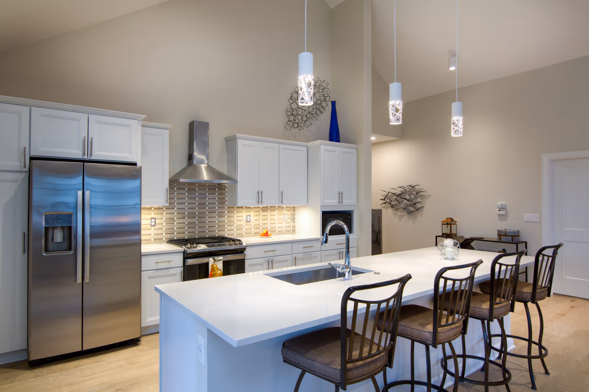 Modern kitchen with white cabinets, stainless steel refrigerator and stove, a large white island with a built-in sink, and four high chairs. Three pendant lights hang above the island, and decorative wall art is visible in the background.